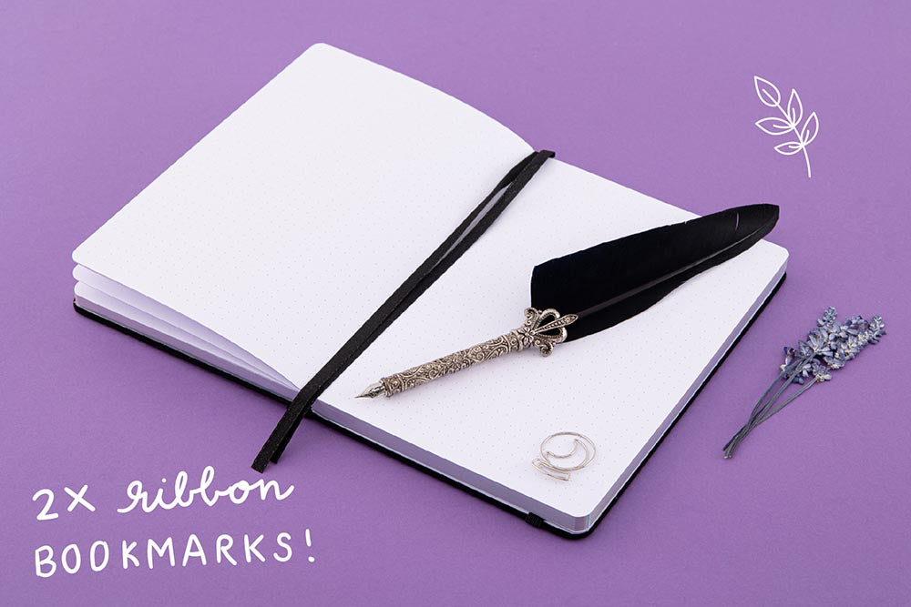 Open page spread of Tsuki ‘Moonlit Spell’ Limited Edition Holographic Bullet Journal with two bookmark ribbons and free bookmark gift with black feather quill and lavender sprigs on purple background