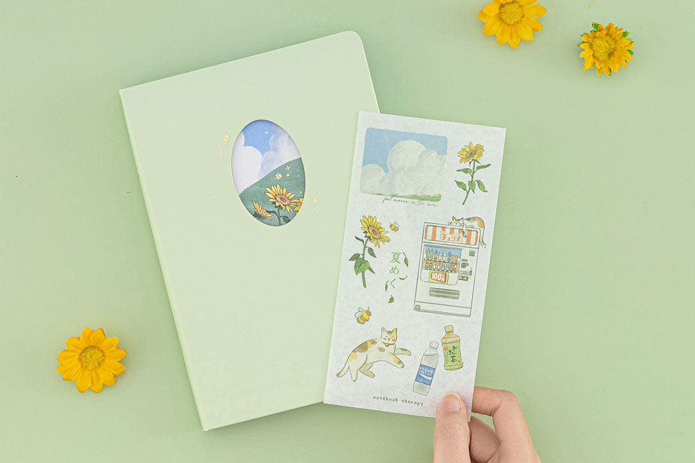 Tsuki Four Seasons Summer Collectors Edition 2022 sage bullet journal notebook on sage green background with sunflower decorations and a hand holding the free sticker sheet