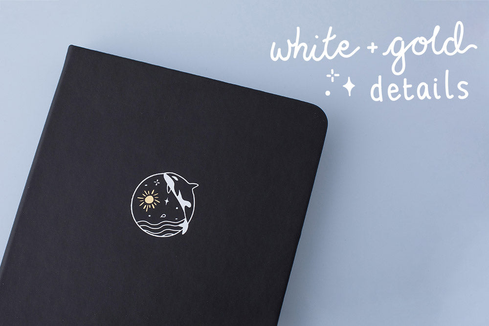 Close up of the front cover of deep black vegan leather Tsuki Playful Orca limited edition bullet journal with white and gold details on blue background