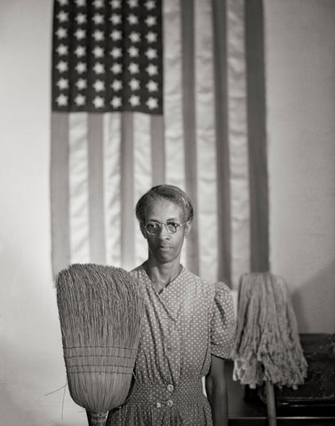 A black woman holding a broom in one hand and a mop in the other. Behind her is the American Flag.