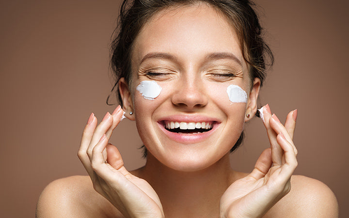 Laughing girl applying moisturizing cream on her face.