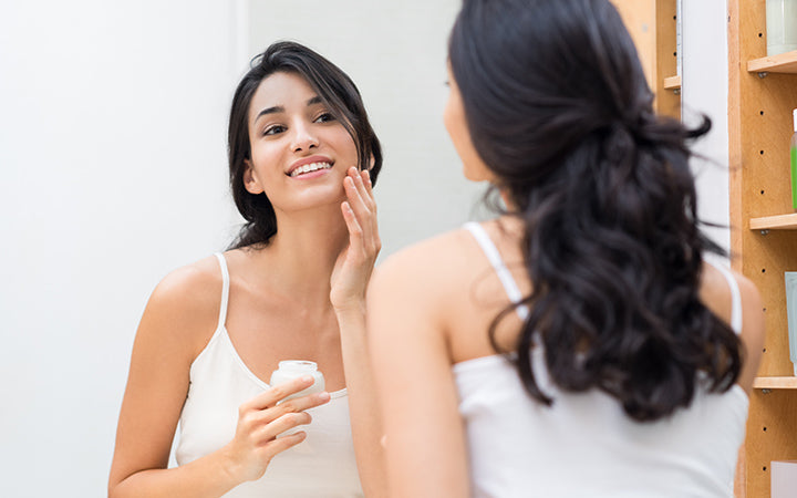 Woman caring of her beautiful skin on the face standing near mirror applying moisturizer on her face