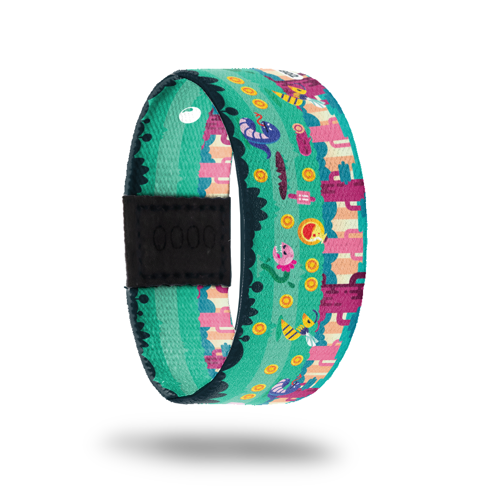 This is a reversible strap. The design is a pixel cartoon of a video game with worms, coins, flower monsters, bees and cacti. The colors are pinks, yellow, blue and green. The inside is the same design and reads Change The Game. It comes with a matching lapel pin and a collector's box. 