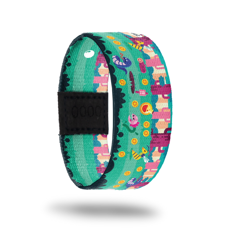 This is a reversible strap. The design is a pixel cartoon of a video game with worms, coins, flower monsters, bees and cacti. The colors are pinks, yellow, blue and green. The inside is the same design and reads Change The Game. It comes with a matching lapel pin and a collector's box. 