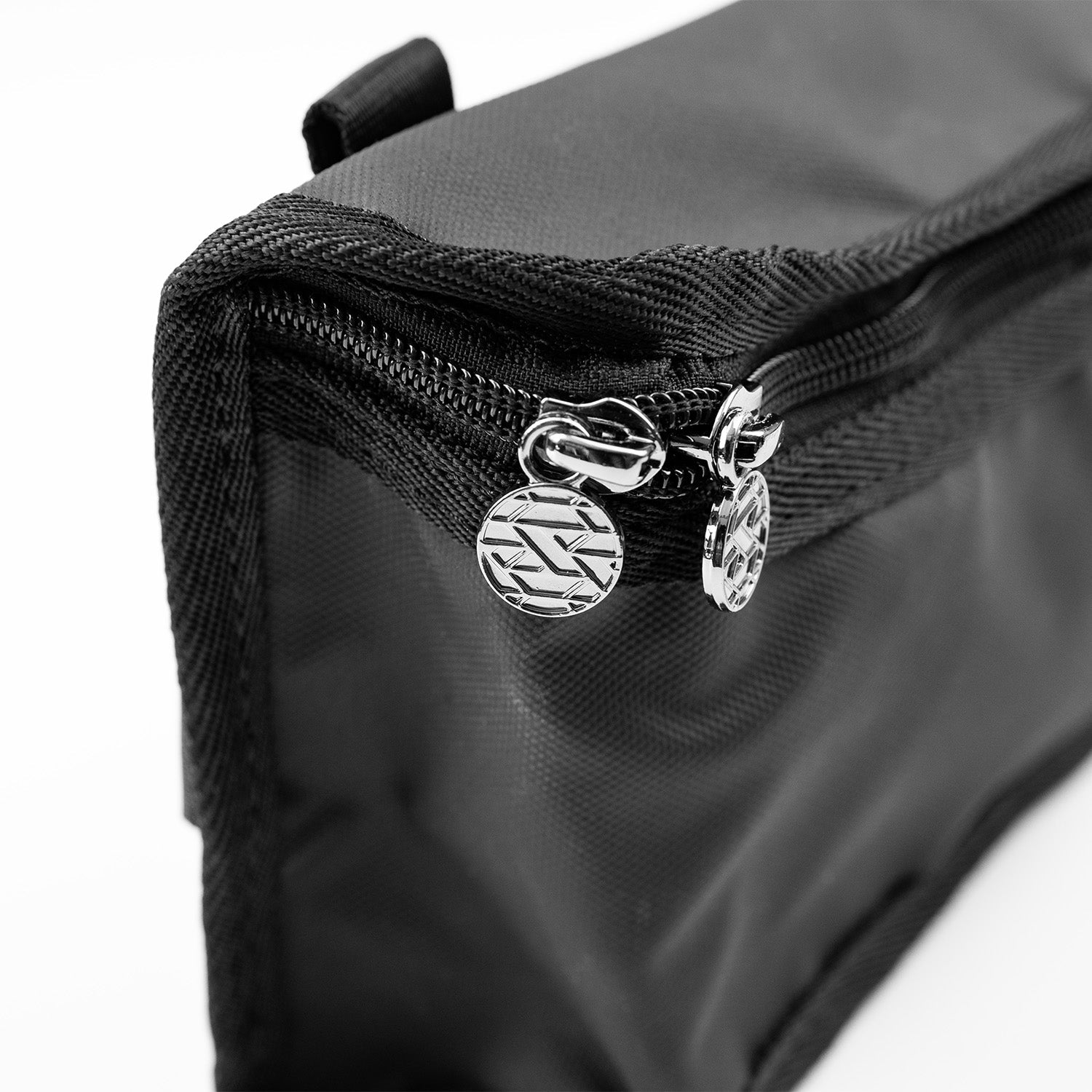 A close-up view of a black waist bag showing the corner of the bag with two silver zipper pulls. The zipper pulls are round and feature an embossed pattern of diagonal lines. The material of the bag appears smooth and durable, with detailed stitching along the edges.