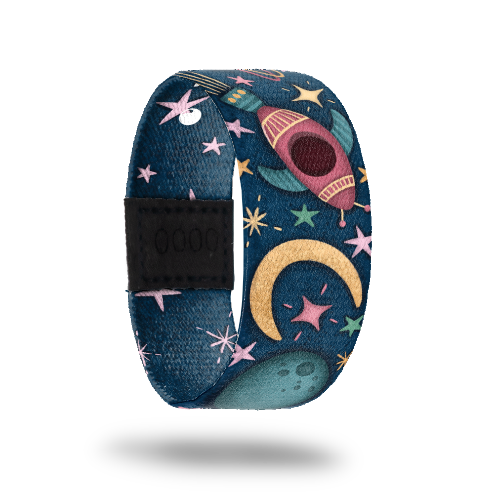 Wristband strap with a dark blue base of space. It has pastel colored stars and bursts with cartoon space ships flying through space. The inside is bluw with all light pink stars and reads One In A Lifetime. This comes with a matching lapel pin and a collector's box. 
