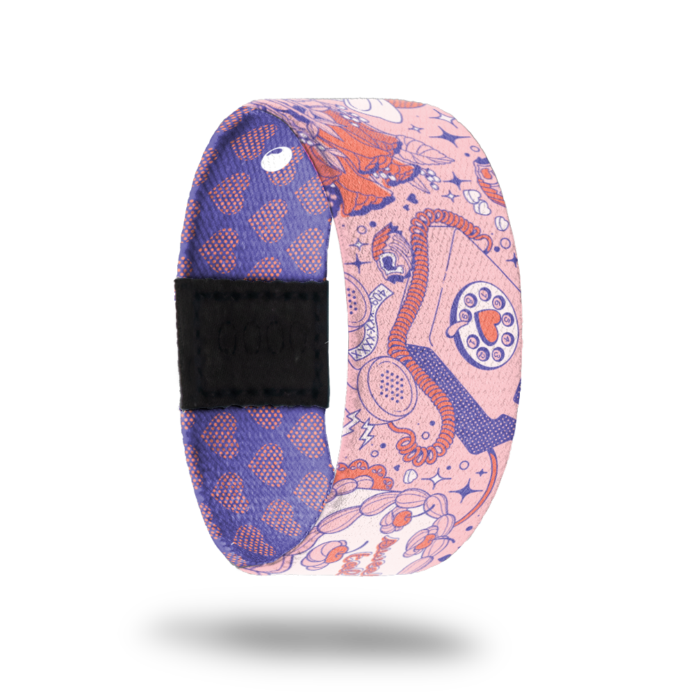 Wristband strap that is pink with red and purple designs of phones, hearts, flowers and diamonds. Inside is purple with pink hearts and reads Full Of Surprises. Comes with matching lapel pin and collector's box. 