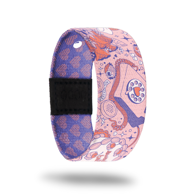 Wristband strap that is pink with red and purple designs of phones, hearts, flowers and diamonds. Inside is purple with pink hearts and reads Full Of Surprises. Comes with matching lapel pin and collector's box. 