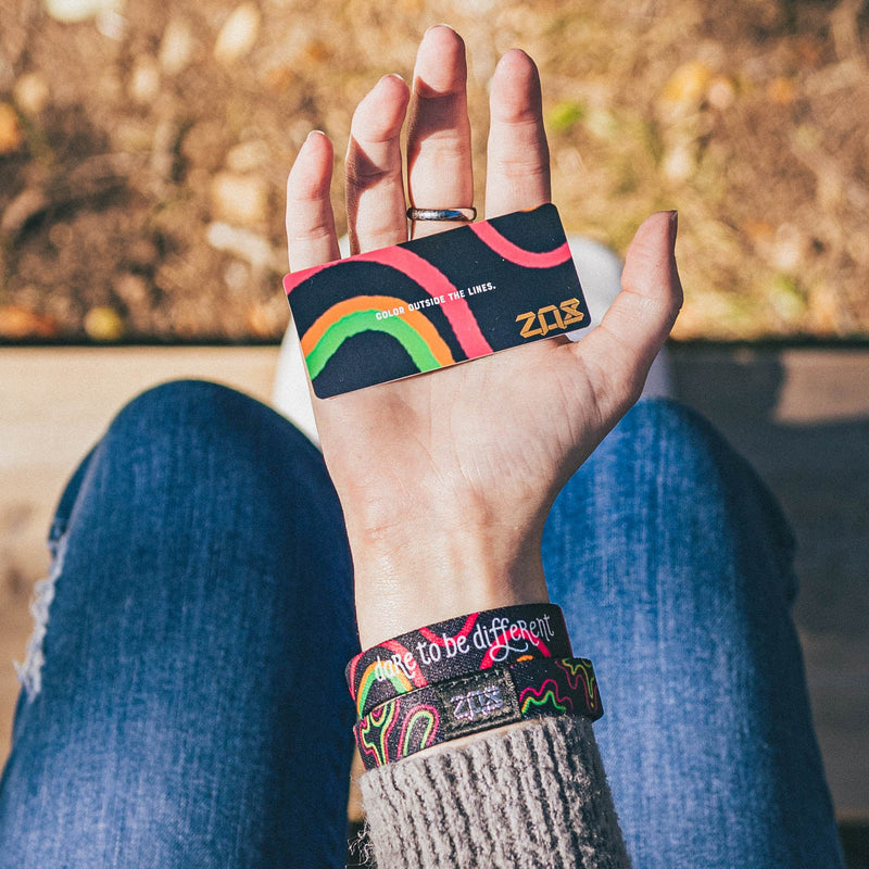 Lifestyle image: hand wearing two Dare to Be Different singles showing back collector's card image of Dare to Be Different: black background with pink, orange and green in zoomed in irregular shapes throughout the single and text ‘Color outside the lines’