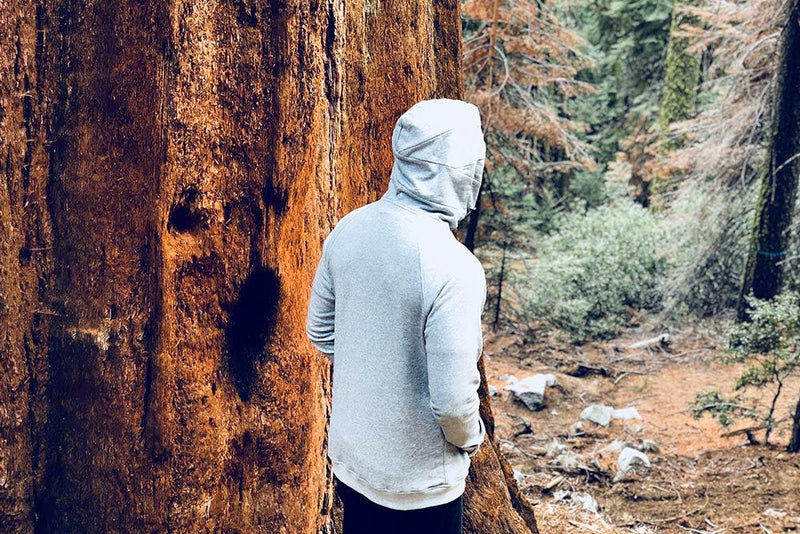 Young Male from the back with his hood up wearing a grey imperial zip hoodie while walking in a forest and is next to a large tree