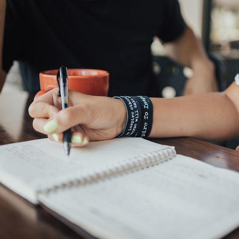 Lifestyle image close up of hand writing in a notebook with 2 Aspire To Inspire on their wrist