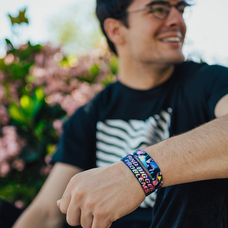 Lifestyle photo of wrist up close of guy wearing two find bigger problems singles one showing the inside design with bold text find bigger problems in a pink to yellow gradient with transparent neon geometric designs overlaying a black background below the single showing the outside design of find bigger problems with neon geometric design overlaying a black background