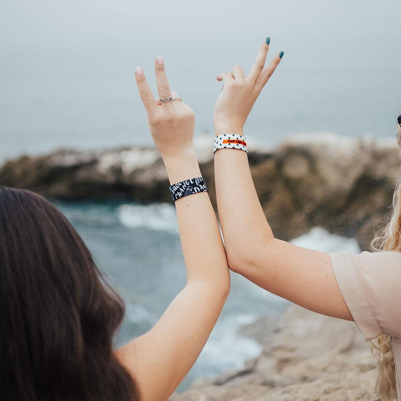 Lifestyle image of two arms raised up next to each other giving the peace sign with hands while each wearing a Good Vibes Only strap