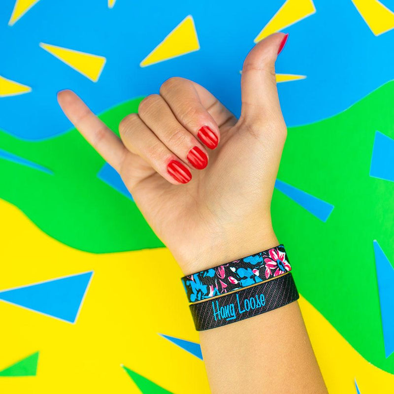 Studio photo of hand signing hang loose showing wrist wearing two hang loose singles one with the outside design of neon tropical flower print overlaying black background and above the other showing the inside design of hang loose with italic text hang loose with grey and black striped background