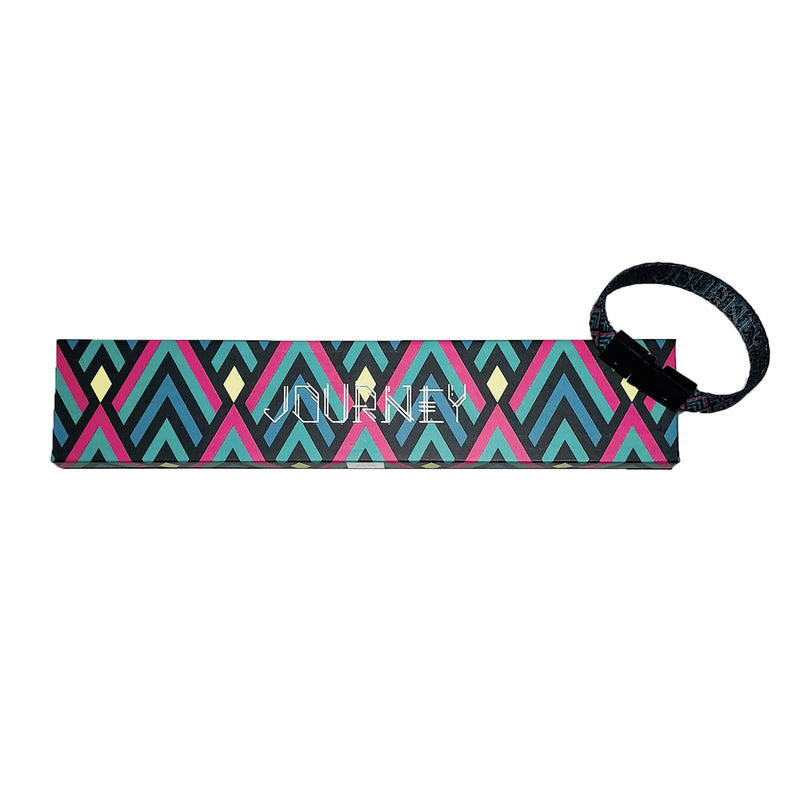  Studio image of Journey clasped together laying on the box it comes in, which has a design of layered triangles with lines of black, pink, and teal in them and Journey centered in whitel text.