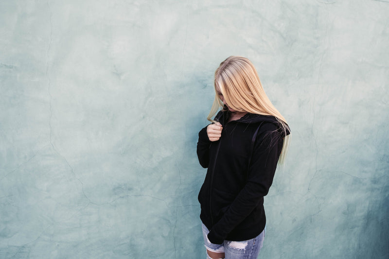 A young adult female zipping up her black imperial hoodie while standing next to a solid light green wall