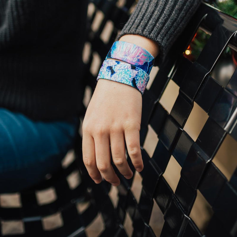 Lifestyle close up image of arm resting on a bench and wearing two Kindness Is Contagious straps