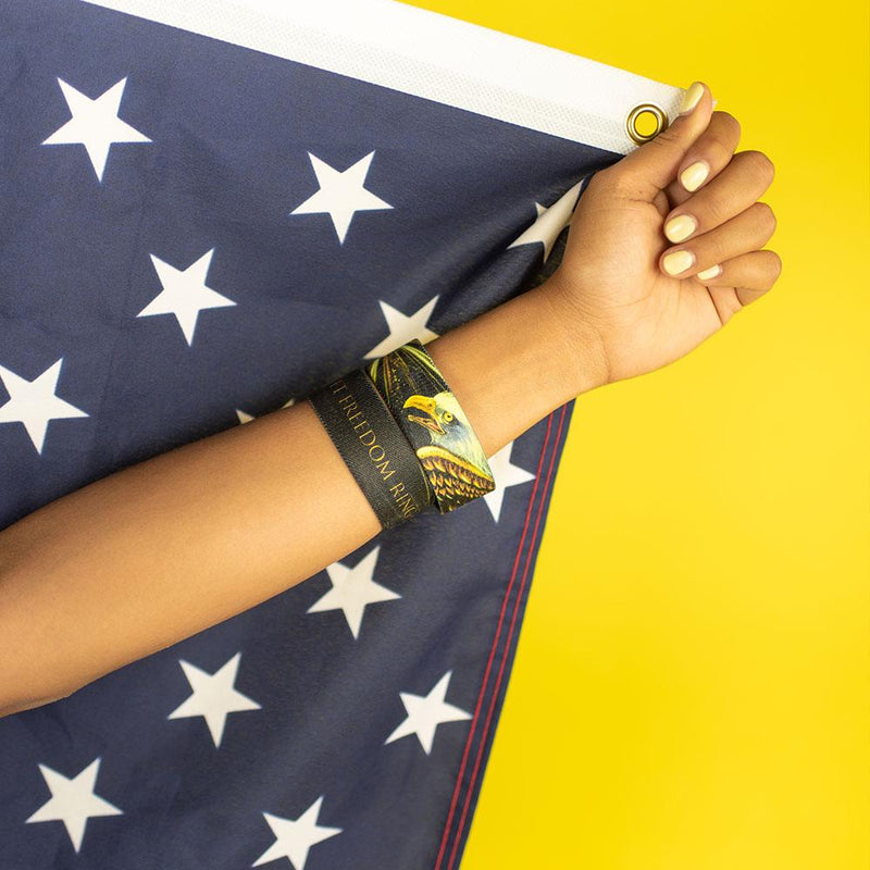 Studio close up of model holding part of a flag with a blue background and white stars while wearing 2 Let Freedom Ring on their wrist