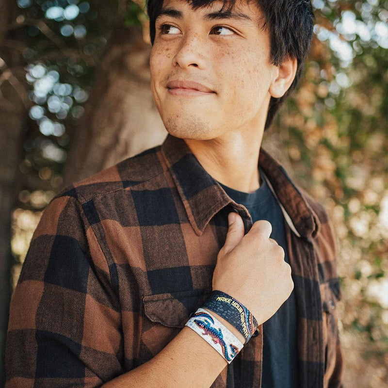 lifestyle image of a man holding his plaid shirt collar wearing two Make your own path wristbands. One showing the inside design and the other showing the outside.