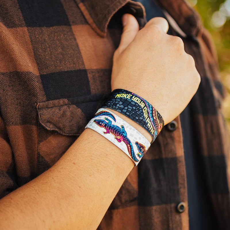 lifestyle image of a hand holding a plaid shirt wearing two Make your own path wristbands. One showing the inside design and the other showing the outside.