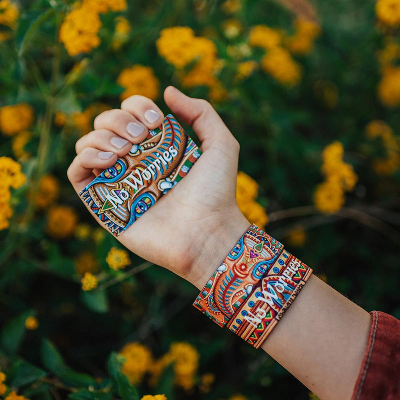 Lifestyle image close up of a hand holding a colorful card that say No Worries in white text and two No Worries straps on their wrist