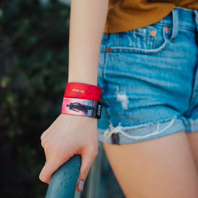 A lifestyle photo of a woman wearing two Show Up wristbands at the same time. One showing the inside and the other showing the outside while her hand is holding a rail