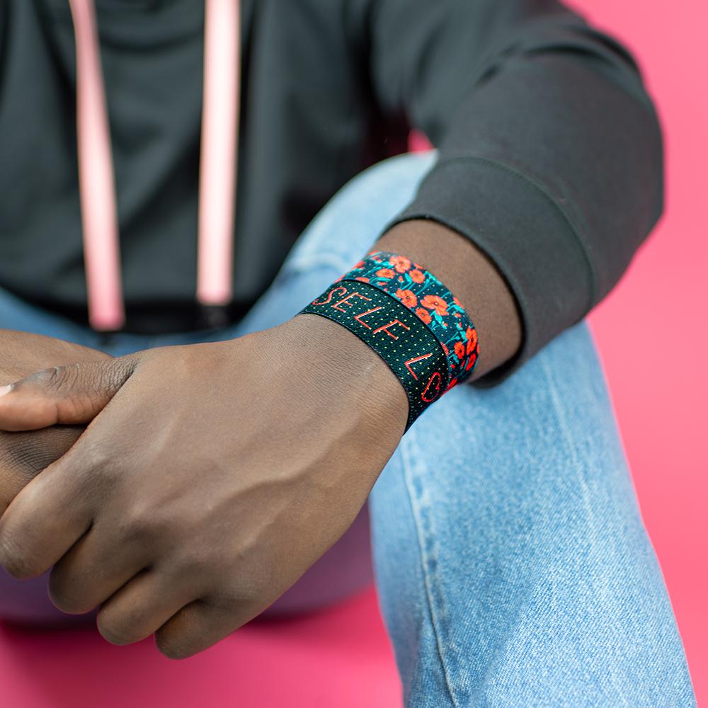 Studio image of sitting model with arms crossed in front of their knees and 2 Self Love on their wrist