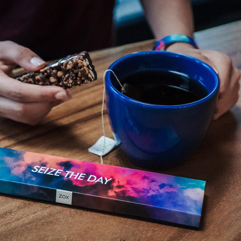 Lifestyle close up image of someone having tea and a biscuit wearing Seize The Day with it's box on the table next to the cup