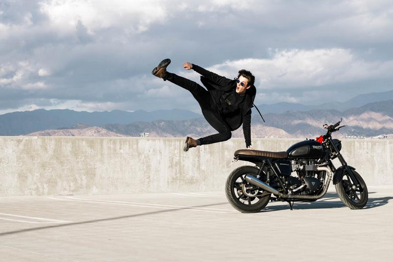 a young male jumping and kicking in the air while next to his motorcycle while wearing a black zipper imperial hoodie