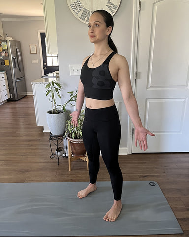 Daniela Spear doing a Standing Spinal Wave with legs shoulder width apart and palms outstretched facing forward