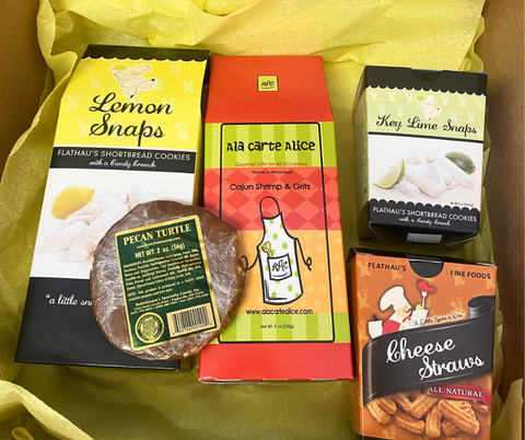 Gift box with yellow tissue paper holding five items: 8oz Lemon Snaps, 4oz Key Lime Snaps, 3.5oz Cheese Straws, 2oz Pecan Turtle, and a box of Shrimp and Grits mix from the company A La Carte Alice