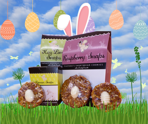 Background of tall, green grass in front of a blue sky with clouds; Boxes of 8oz Key Lime Snap cookie carton, 6oz Raspberry Snap cookie carton, 4oz Lemon Snap cookie carton and 3 chocolate pecan turtles with white chocolate easter eggs sitting in front of the cookie cartons in the grass; Key Lime and Raspberry box have rabbit ears on top, as if they're wearing the ears; A string of Easter eggs hangs from the top of the graphic and seen behind the products in the sky;