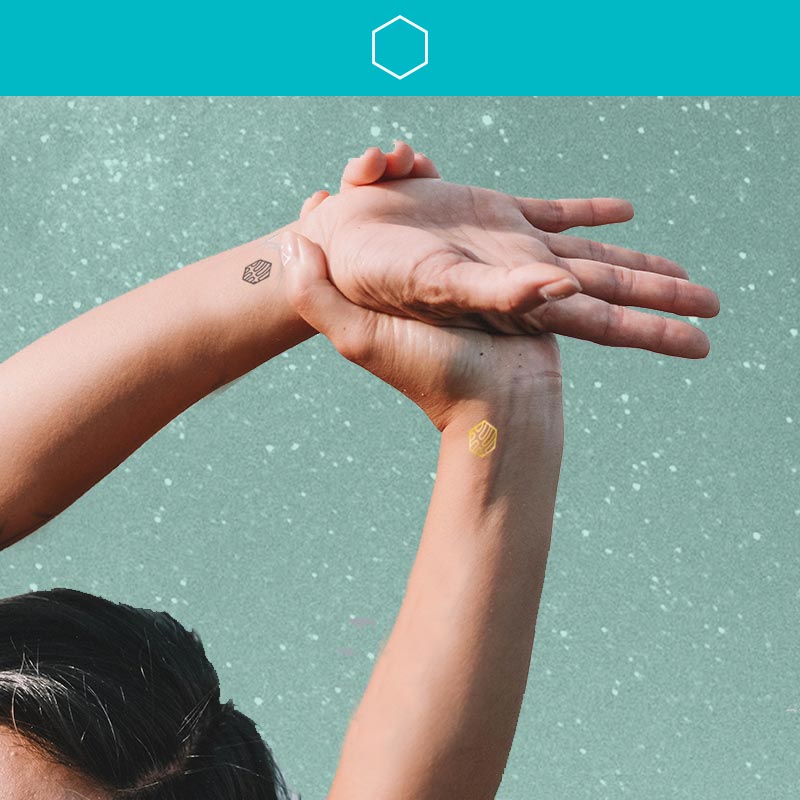 Top of person's head with streak of grey hair reaching arms over head and holding wrist that have mindfulness temporary tattoos that represent kindness.