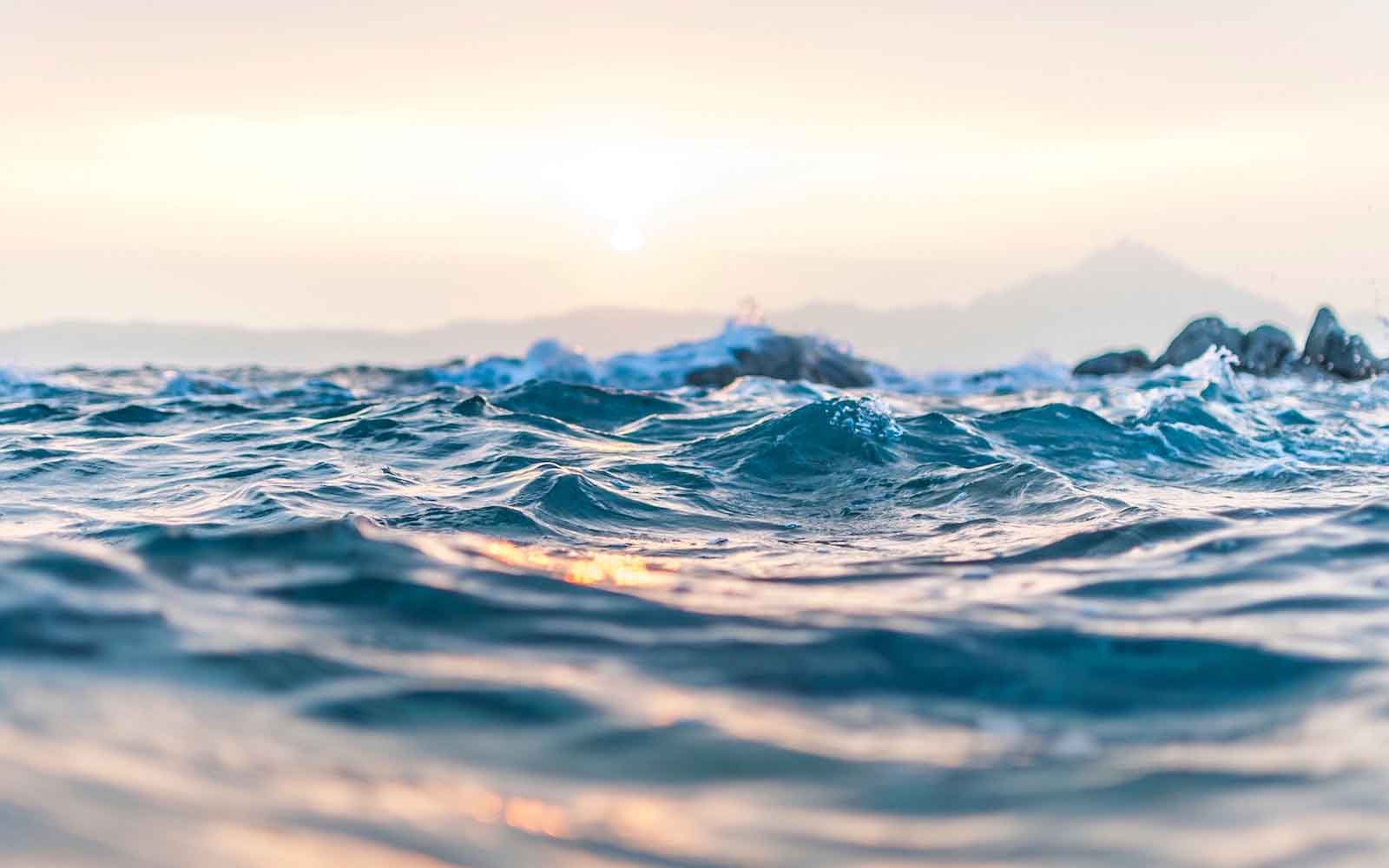 Close-up of waves splashing with mountains and hazy sunset in background.