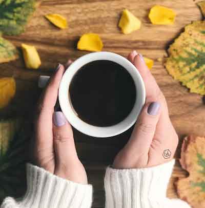 Girl wearing sweater and flow intention tattoo while holding cup of coffee on a table with fall leaves. 