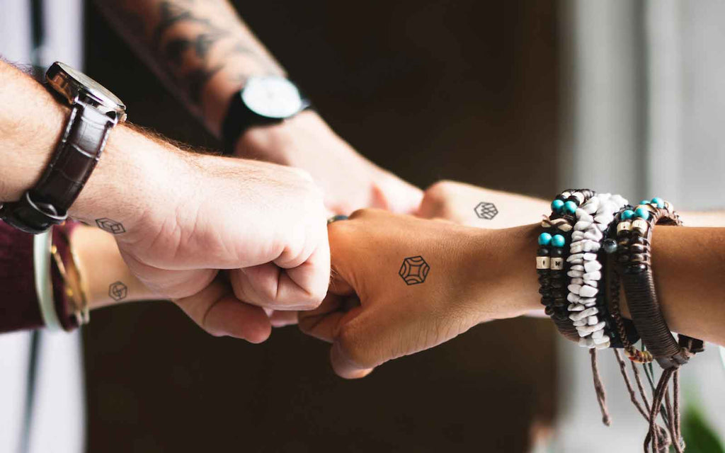 Five hands fist bump. Two hands have half-inch black mindful marks on top of hand. Others have temporary tattoos on wrists.