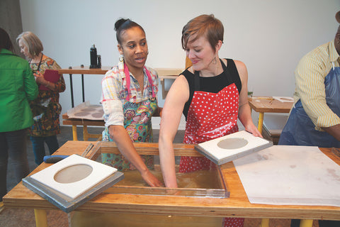 Two women standing over a wood frame table with a tub of water built in. Each has an arm in the tub, as if stirring the water. One is looking at the other smiling while the other looks at the water, also smiling. There are other people in the background and two metal square plates with a white mold with a circle cutout on them. A finished sheet of paper is on the table next to the tub. 