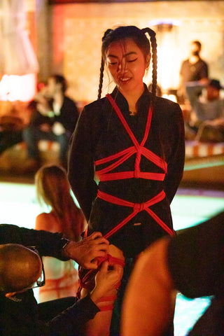 Young woman smiling in black robe being tied with red rope by a shibari master who is kneeling while tying intricate knots on her thigh