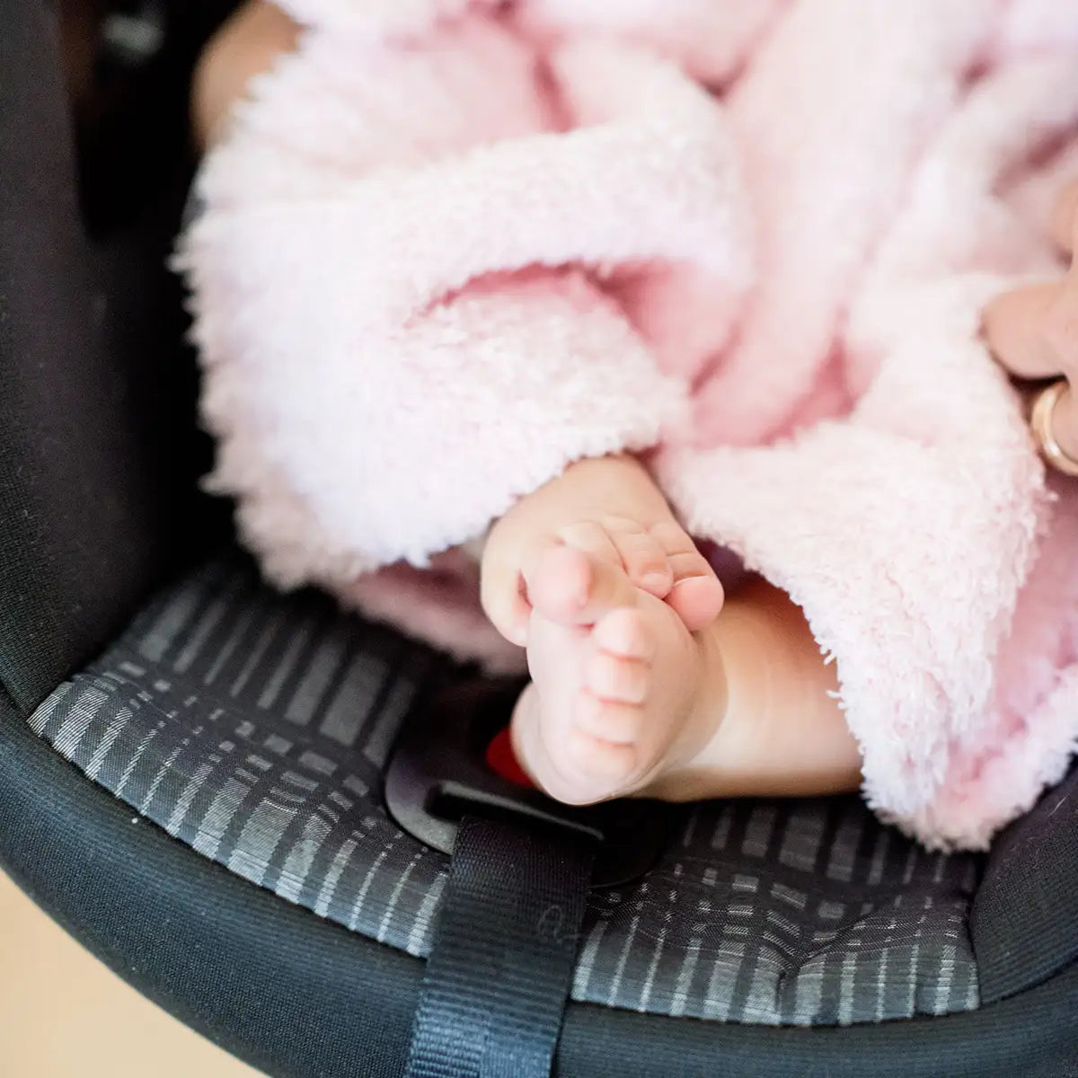 A close-up of a baby's feet resting in a car seat, covered by a soft pink blanket.