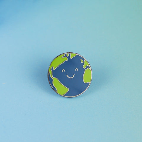 An Earth enamel pin sits on a blue background. The earth design has a cute smiling face.