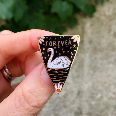 A hand holding an enamel pin with a swan detailed on it. 