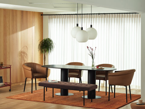 Dining area, large leather armchairs, black wood pedestal dining table, maroon fabric bench, white globe chandelier. Plants. Light walnut wall and white curtains.