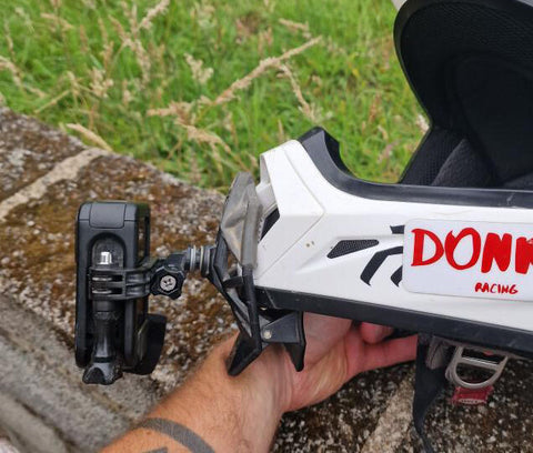 A black action camera is attached to a white motorbike helmet, positioned in a portrait orientation using a clip on mount to the chin area of the helmet and an adapter to position it in the correct way.