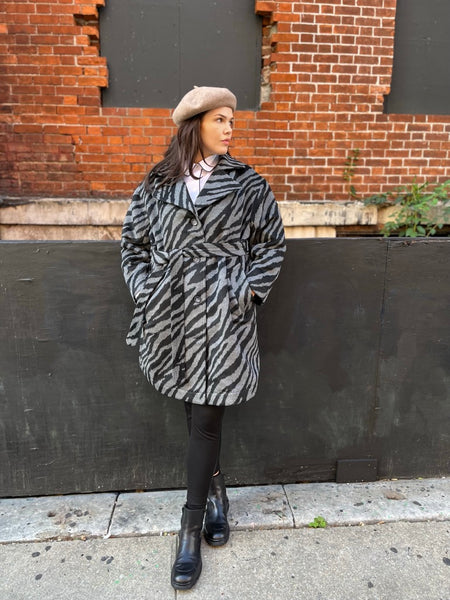 Model wearing See ROSE Go gray and black zebra coat leaning against a red brink wall layered over black leggings and black booties.