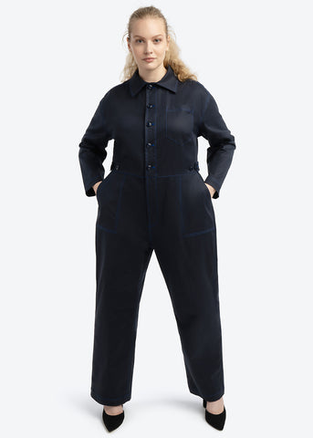 Well heeled, Model is now wearing the same Riveter Jumpsuit in Passport Blue, which can be described as a lighter navy, and wearing heels. Showing the style versatility of the jumpsuit. Models is standing in power pose with hands in pockets and styled with Black Stiletto shoes.