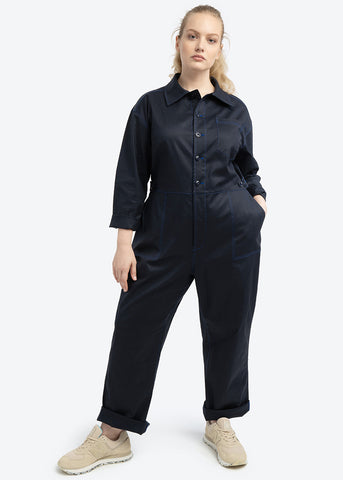 Casual cool, model is wearing the Riveter Jumpsuit in plus size 1x size 14 / 16. Model is posing in a white ground studio with hands in front pockets and wearing neutral beige color sneakers.