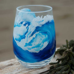 Stemless white wine glass with blue ocean wave painted around the glass, sitting on driftwood at the beach