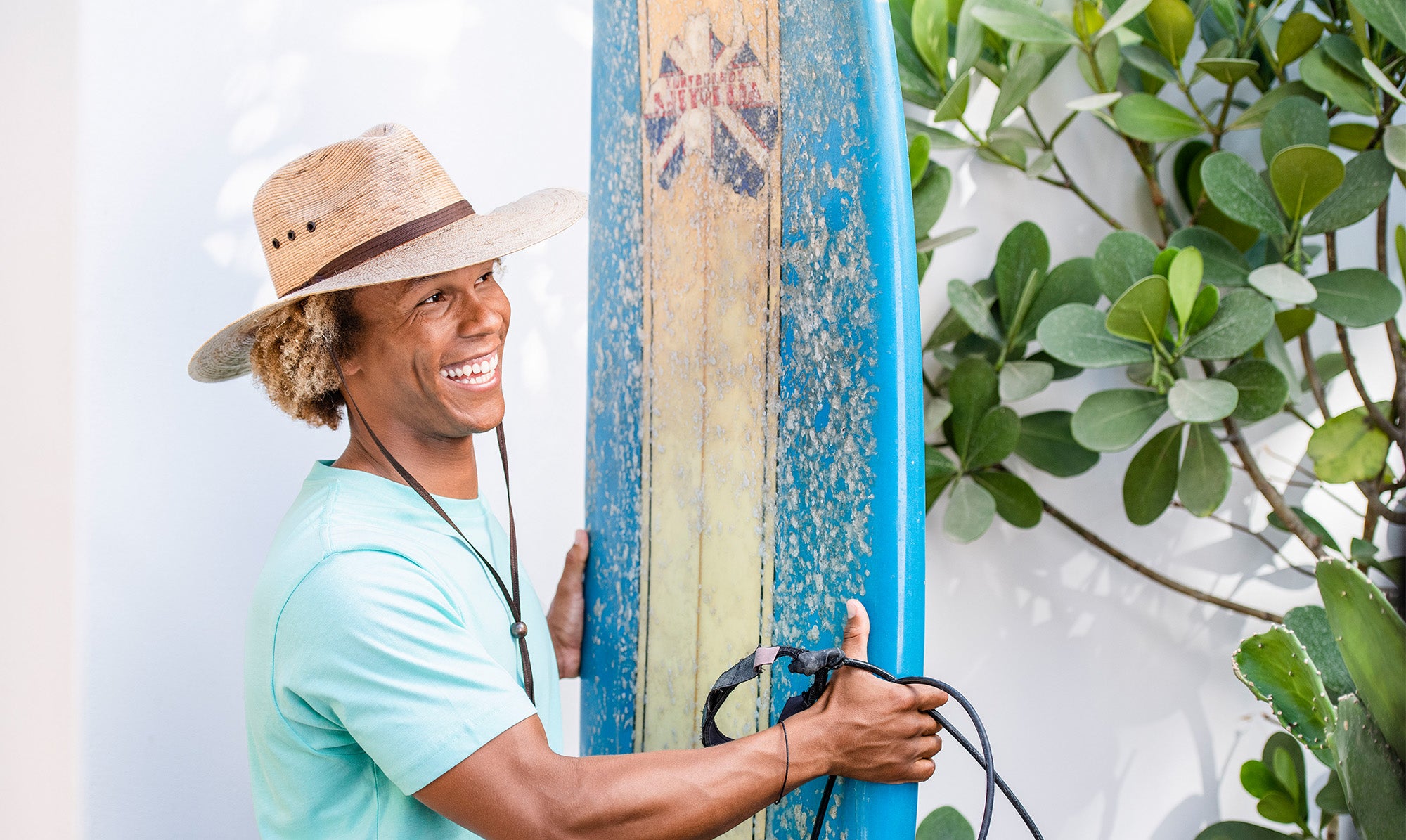 Man Wearing the Wallaroo Big Wide Brim Baja Straw Sun Hat while Standing holding a Surf Board