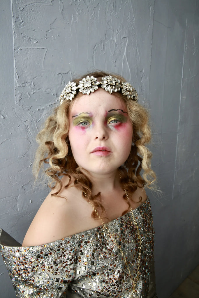 A model with Down Syndrome is wearing a crystal headband by Jolita Jewellery, with the intricate details of the headband visible. The model is smiling and looking confident, with her hair styled in loose waves. The background is blurred, with a soft focus on the model and the headband.
