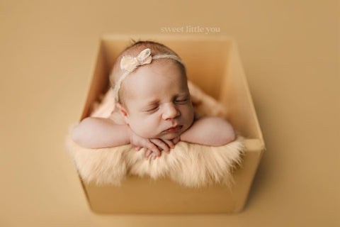 Caucasian baby at a newborn professional photography session. Baby is styled on seamless paper, a wooden crate, faux fur layer, and velvet bow tieback headband all in peachy tones.